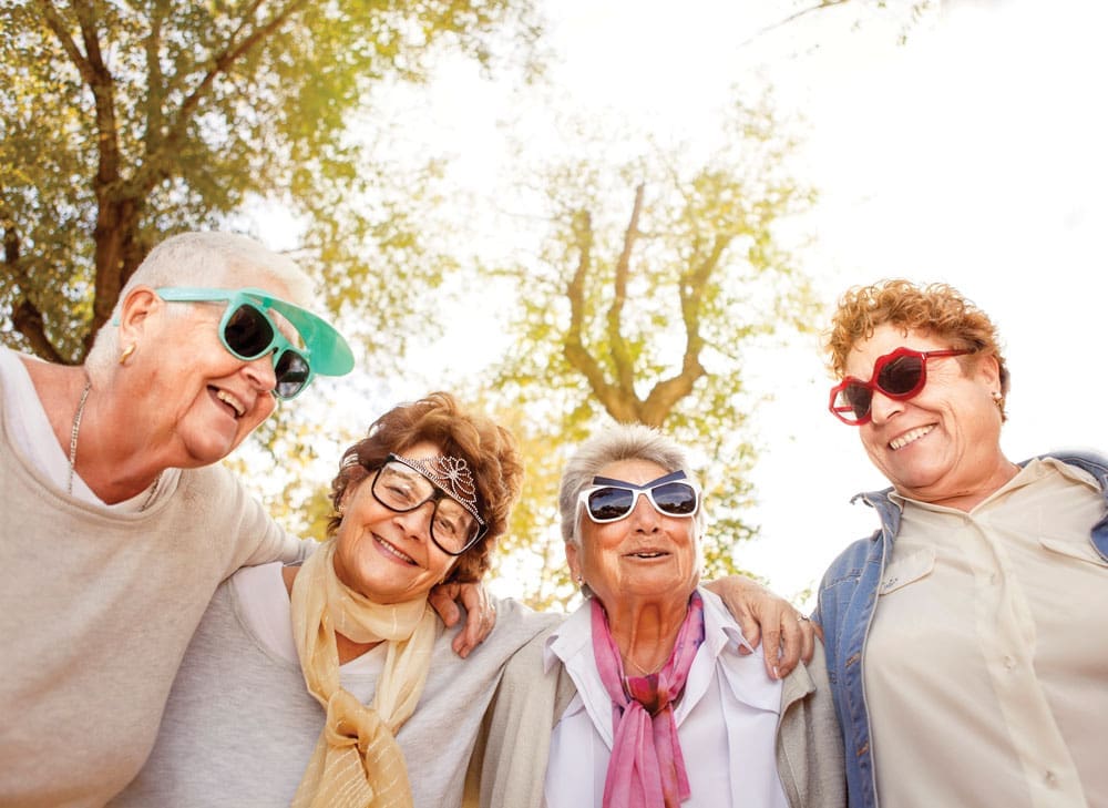 Four older women pose closely together outdoors while wearing funny sunglasses