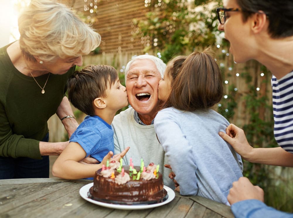 Older man celebrates his birthday outdoors with his family and a cake while receiving kisses on the cheek