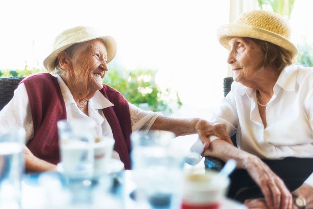 Two older women wearing hats sit next to each other at outdoor table and smile